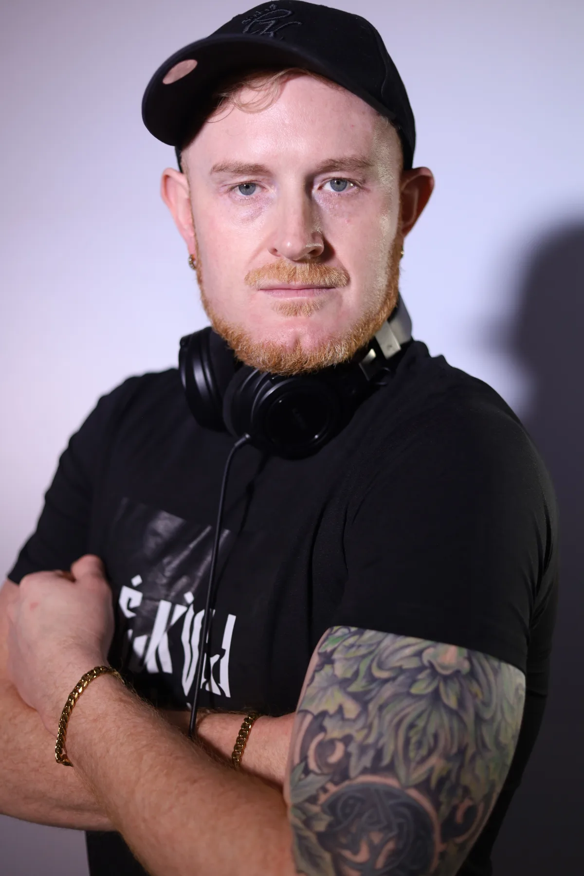 Dave Mladi in a black cap and t-shirt, arms crossed, against a pale backdrop
