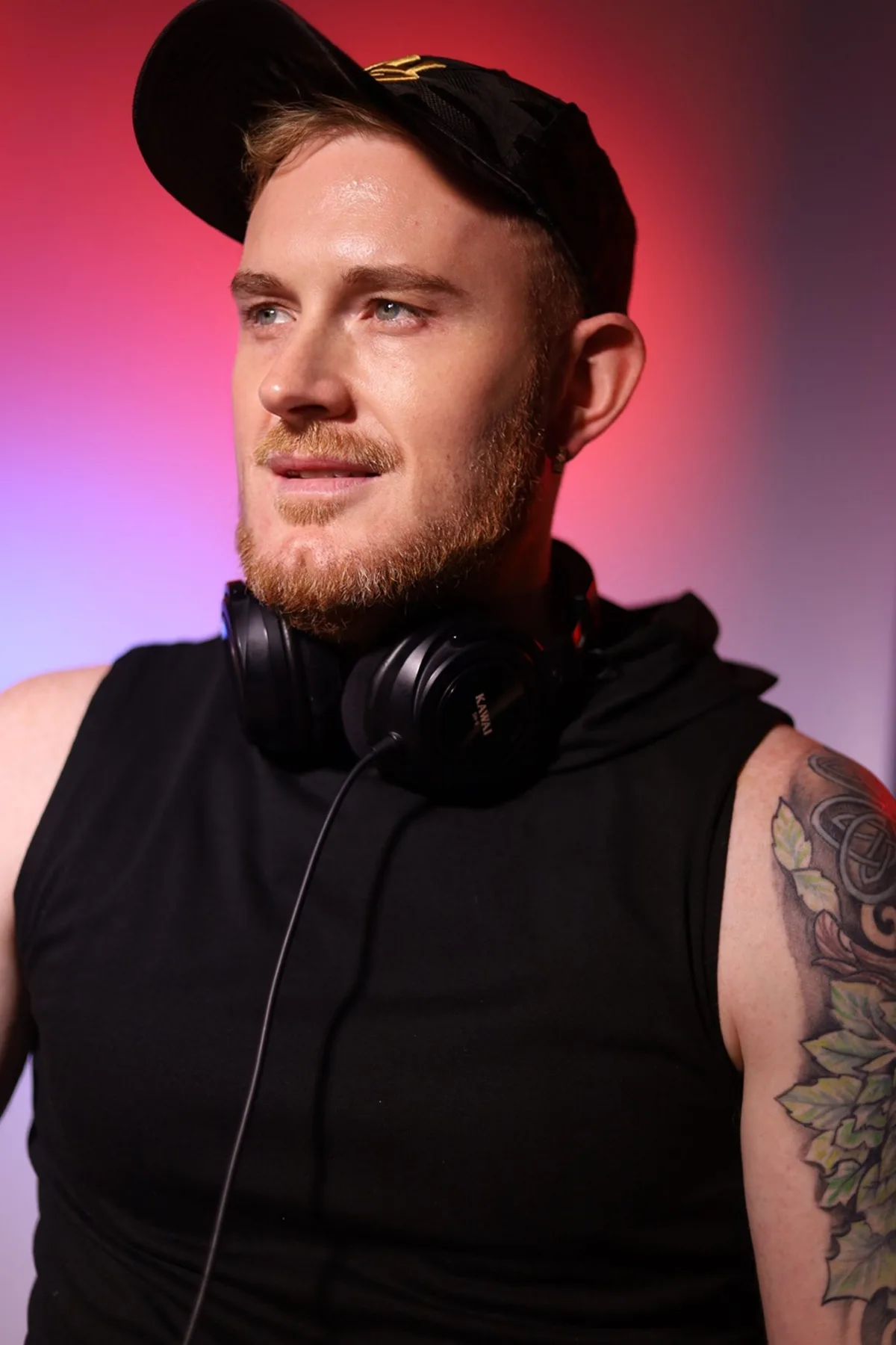 Dave Mladi in a black cap and vest with headphones round his neck, lit by a red backlight