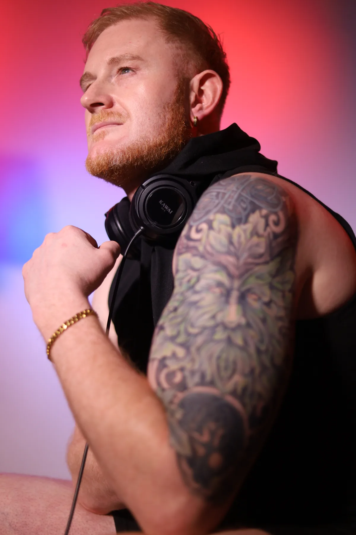 Dave Mladi in profile showing his tattooed arm against a pink and red backdrop