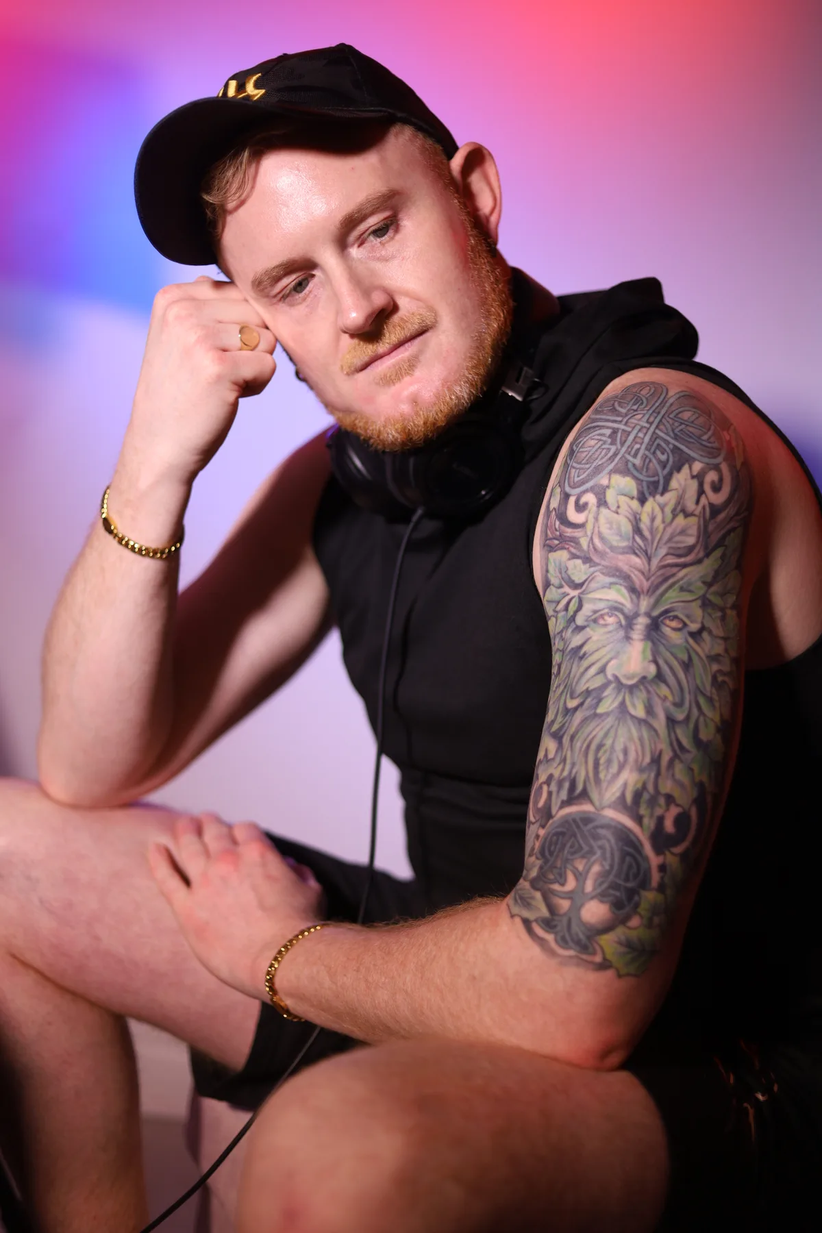 Dave Mladi seated in a black cap, hand to his face, under pink studio light