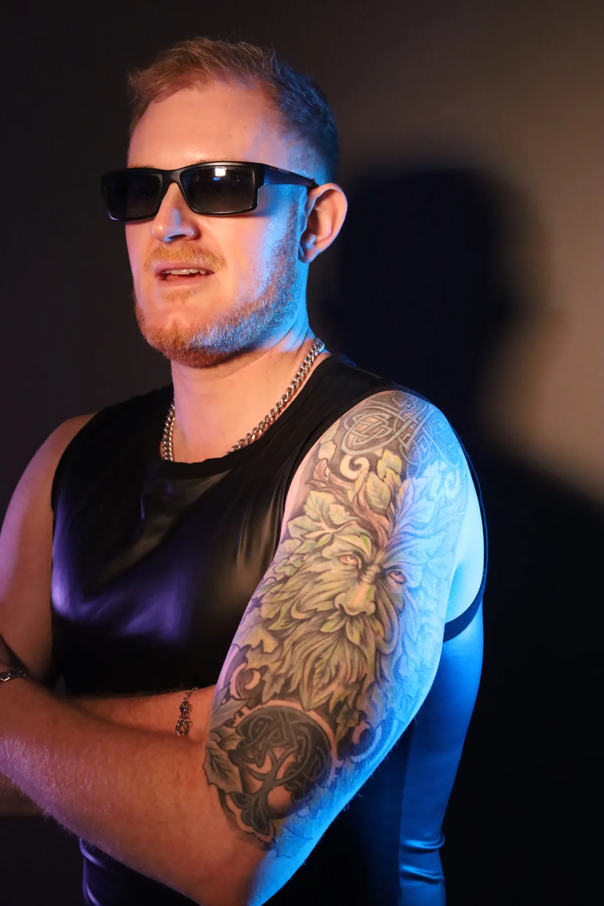 Dave Mladi in sunglasses and a black vest against a dark backdrop