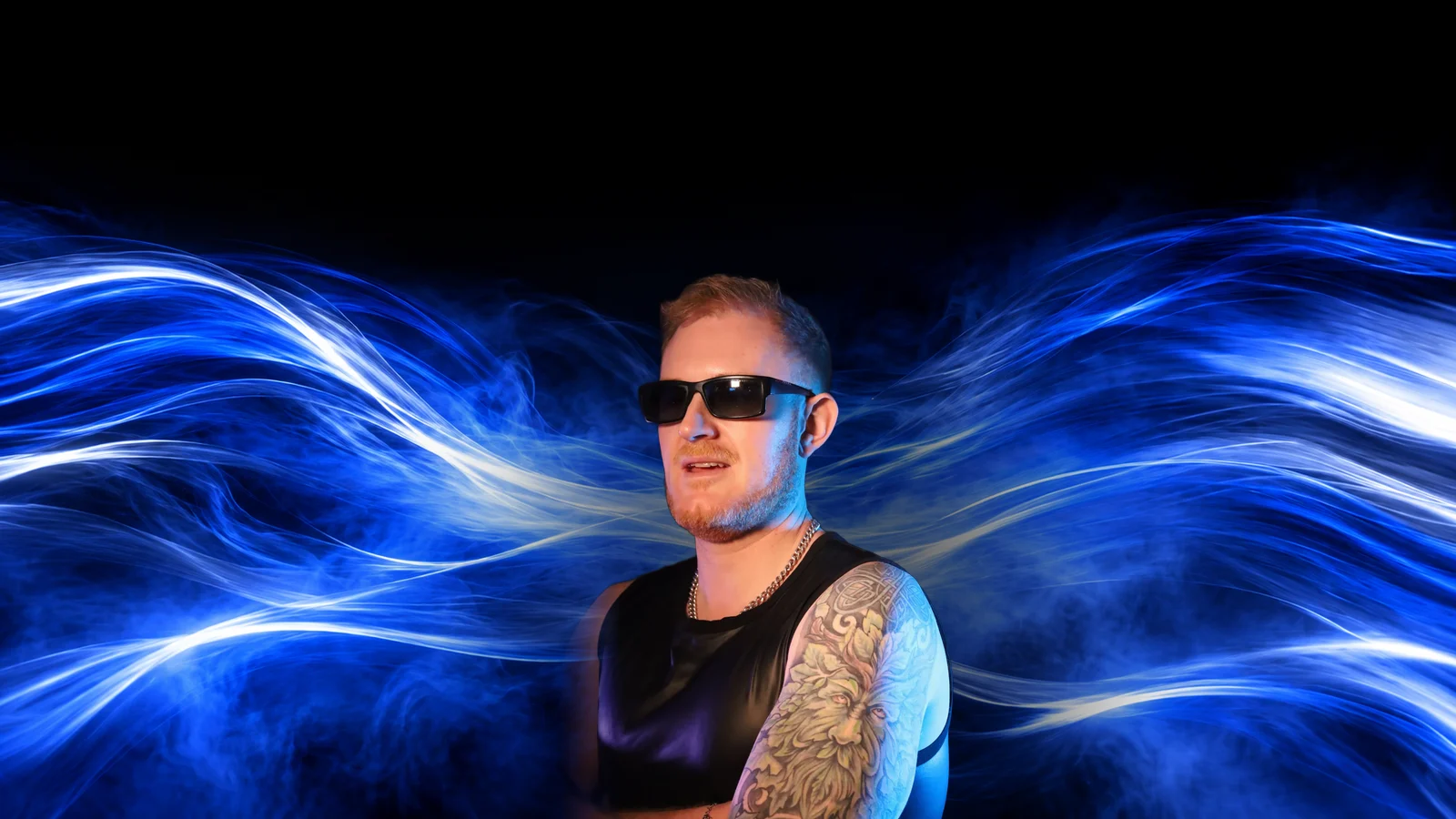 Dave Mladi in sunglasses and a black vest, standing in front of sweeping electric blue light trails